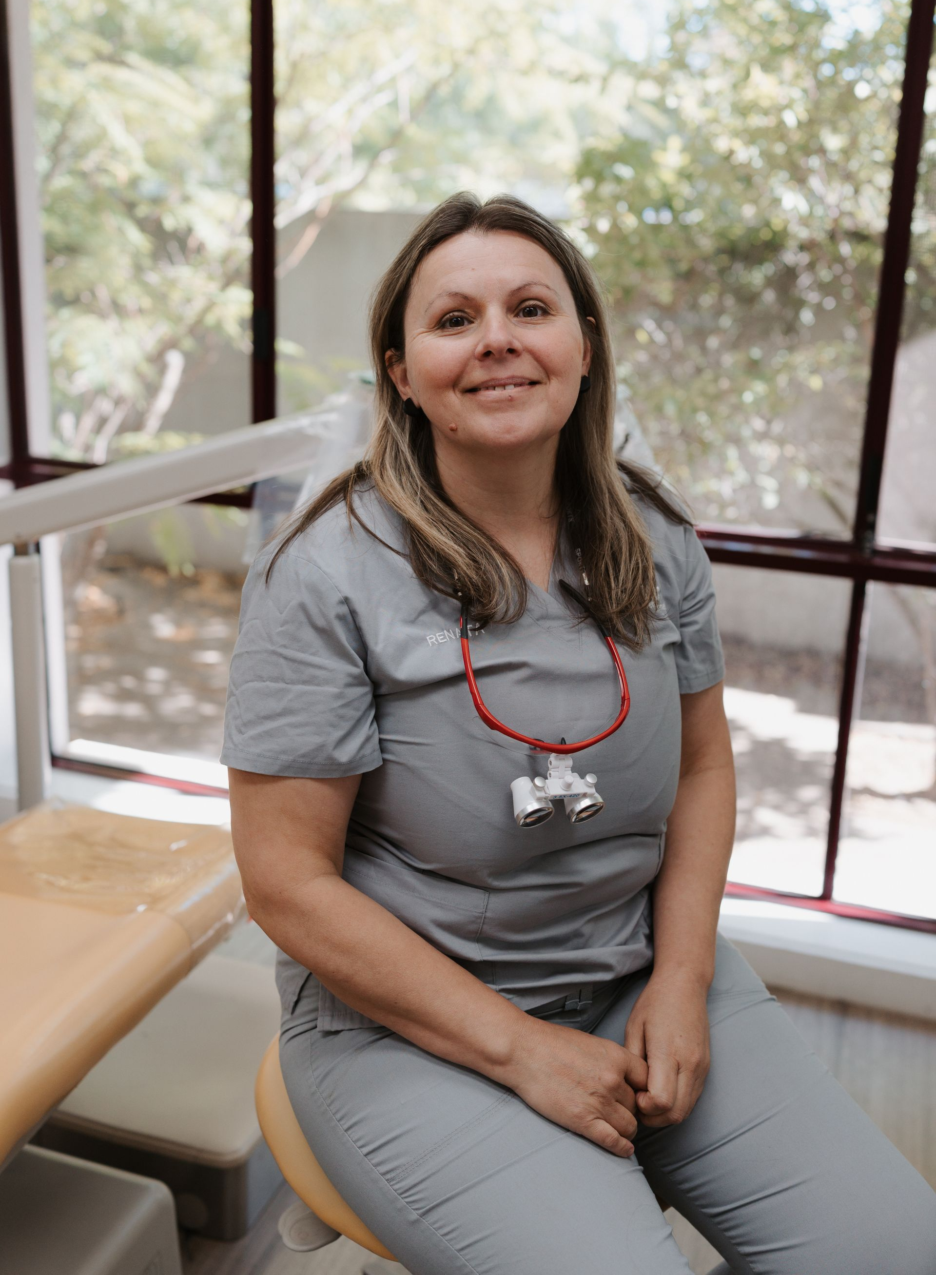 A woman in a grey scrub top is smiling in front of a white wall.