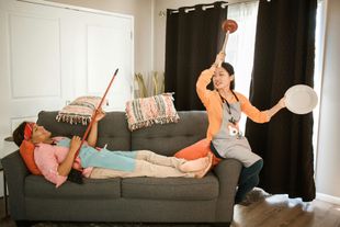A woman is laying on a couch while another woman is holding a plunger and a plate.