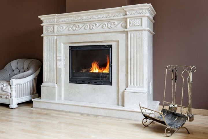 White fireplace with a lit fire in a bright room, beside a chair and fireplace tools on a stand