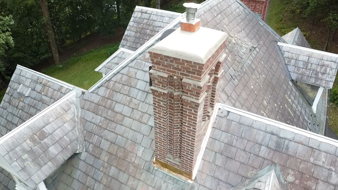 Aerial view of a brick chimney on a gray-shingled roof surrounded by trees.