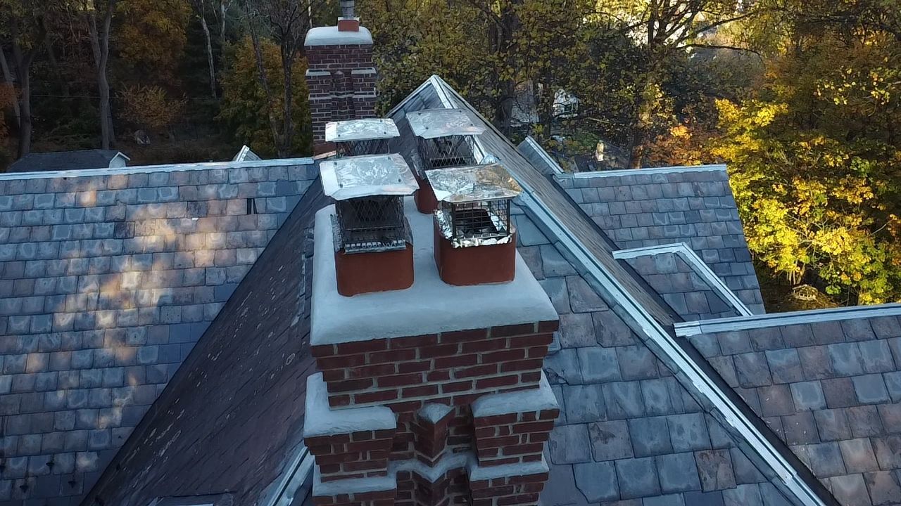 Aerial view of a slate roof with two red brick chimneys and wooded trees in the background