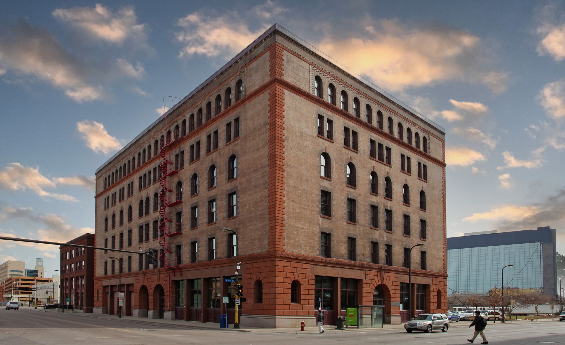 Brick building on a city street, with red trim and fire escape, under a cloudy sky.
