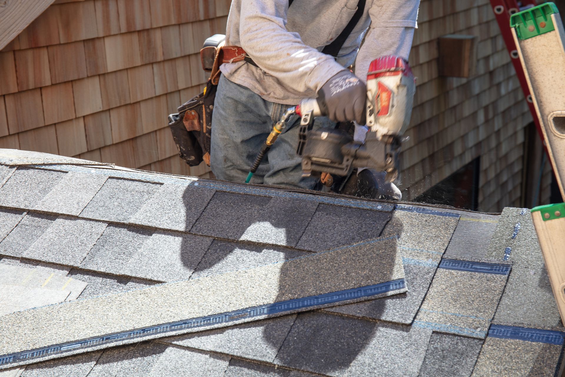 Roofer using a tool on a gray shingled roof, working near a wooden shingle wall and ladder.