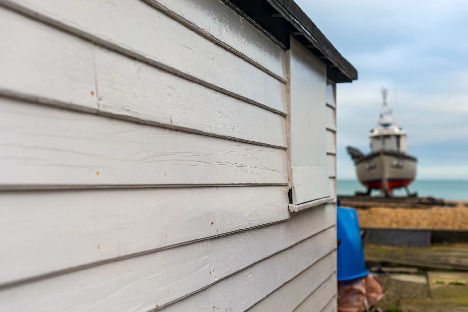 White shed with a blurred boat on the beach in the background. Cloudy sky.