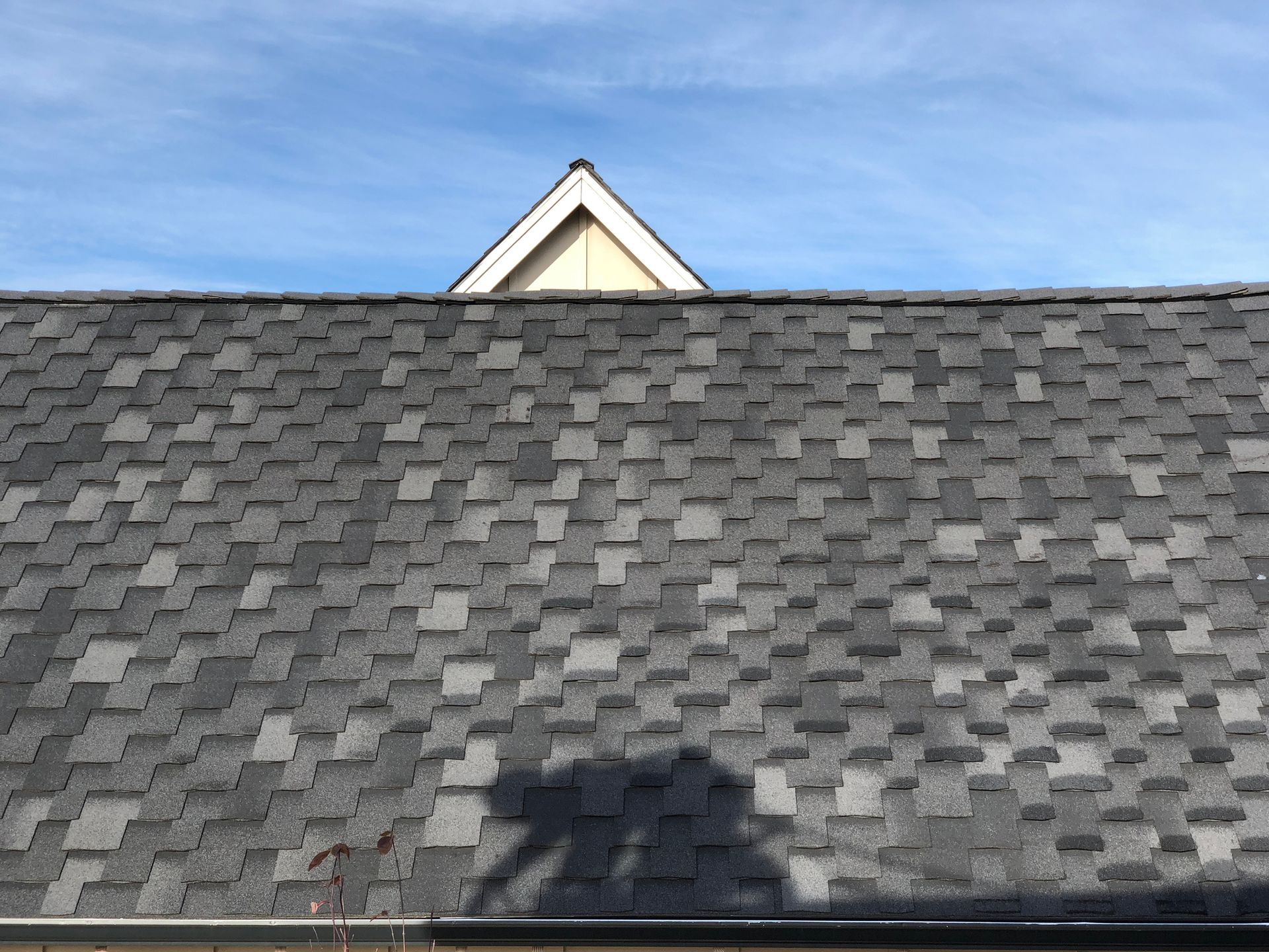 A roof covered in gray and black shingles. In the background, a small triangular roof peak against a blue sky.