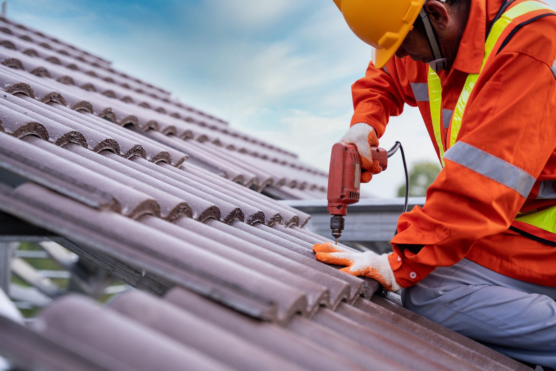 Roofer in orange jacket and hard hat using a drill to install tiles on a roof.