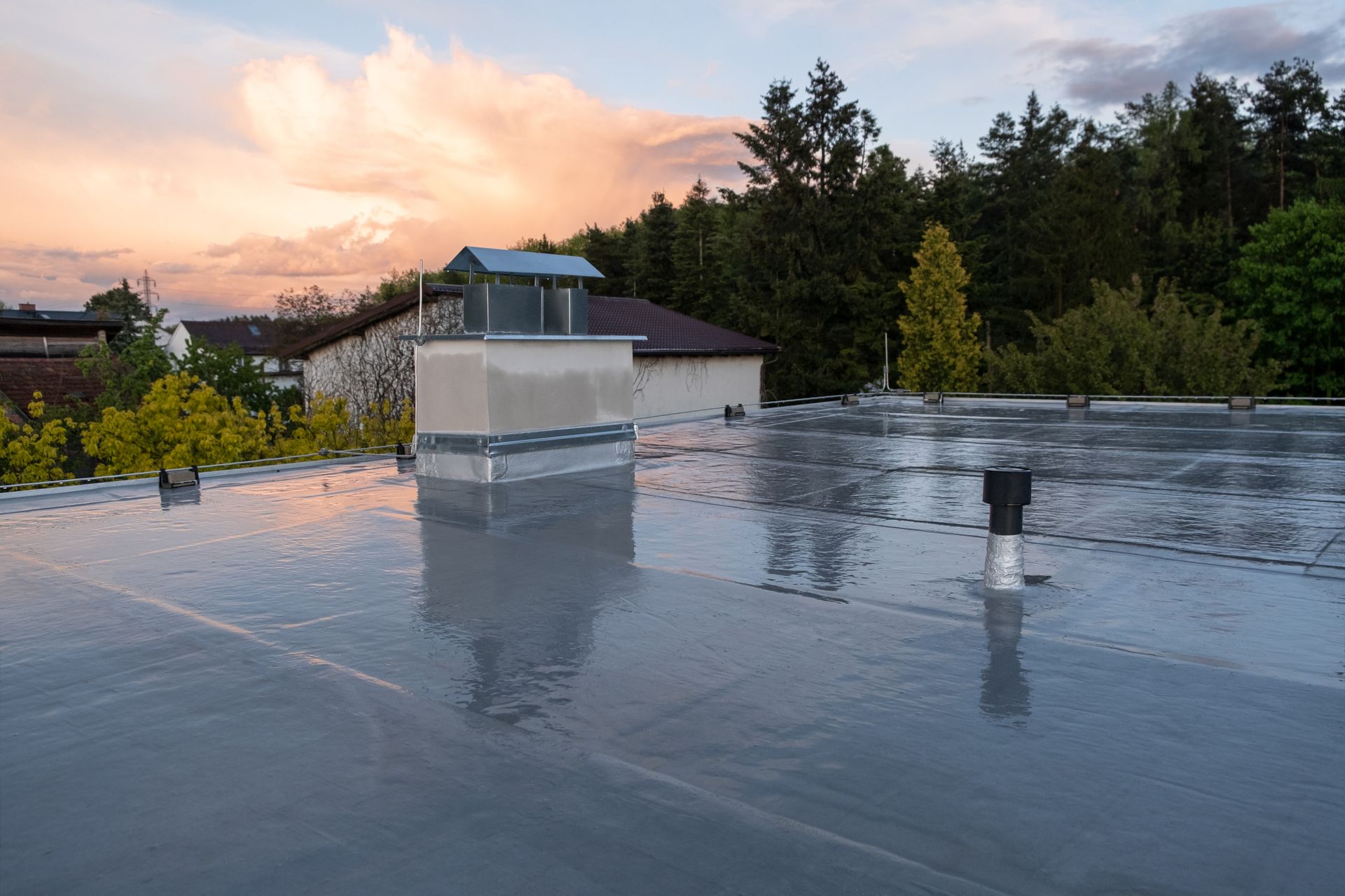 Flat roof with vent, reflections of cloudy sunset, trees in background.