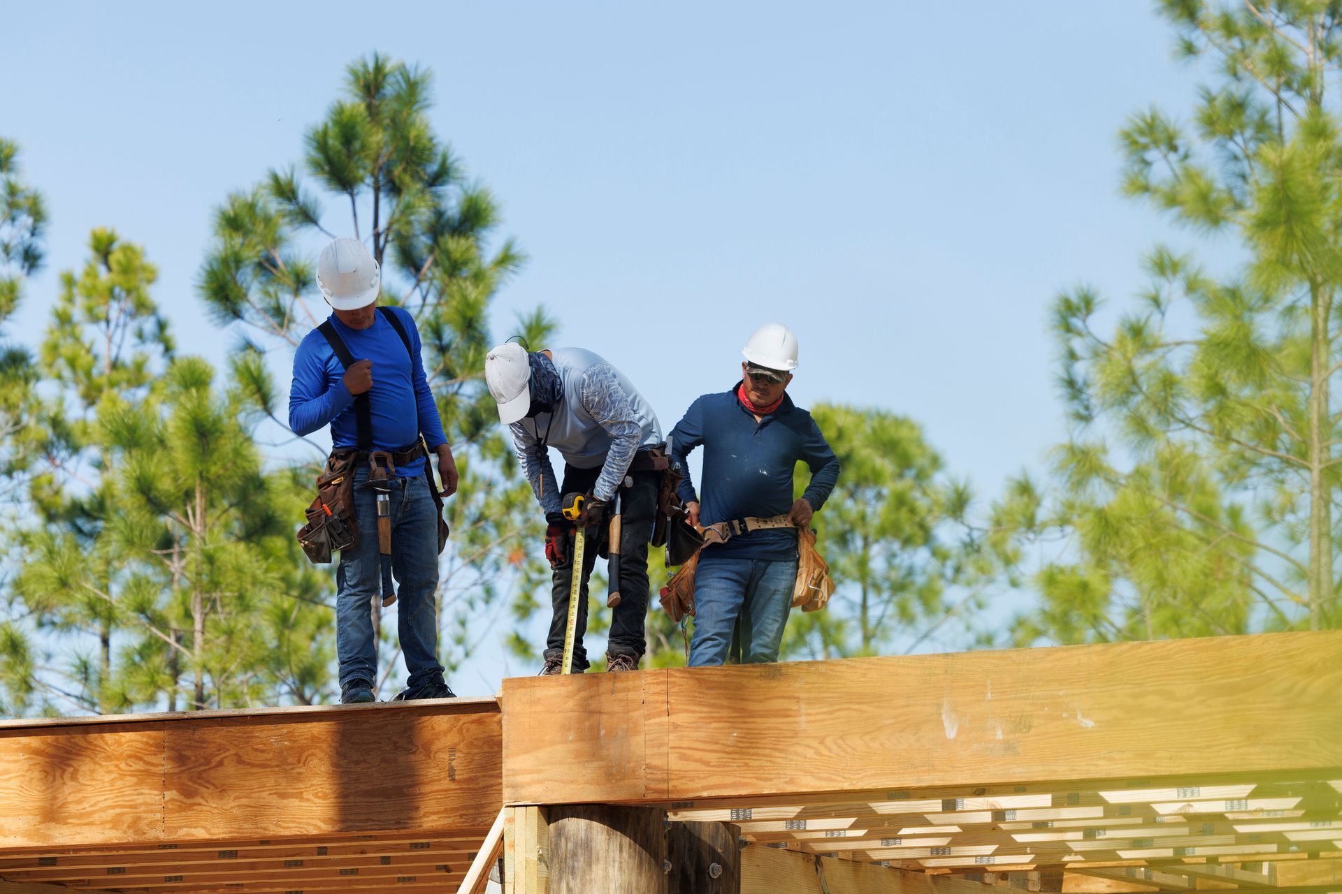 Construction workers on a roof wearing hard hats and safety gear, working on a sunny day.