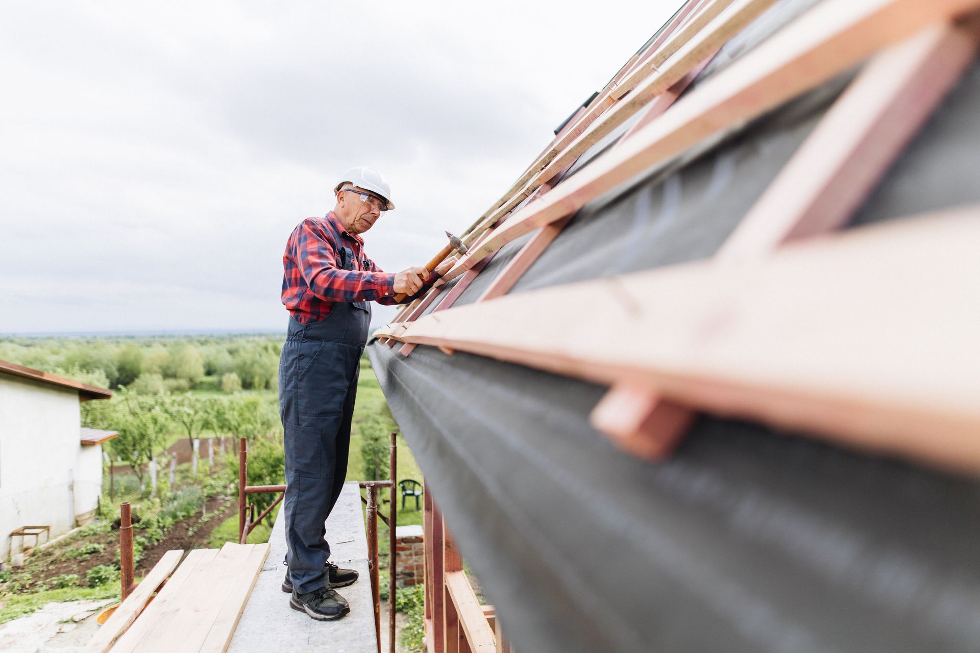 Roofer installing wooden supports on a roof. Outdoors on a cloudy day, he wears a hard hat and overalls.