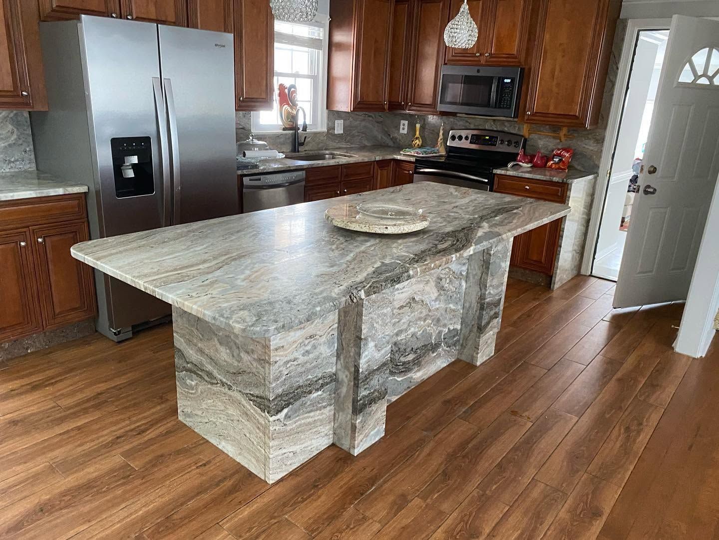 Modern kitchen with a large granite island, wood cabinets, stainless fridge, and hardwood floors.