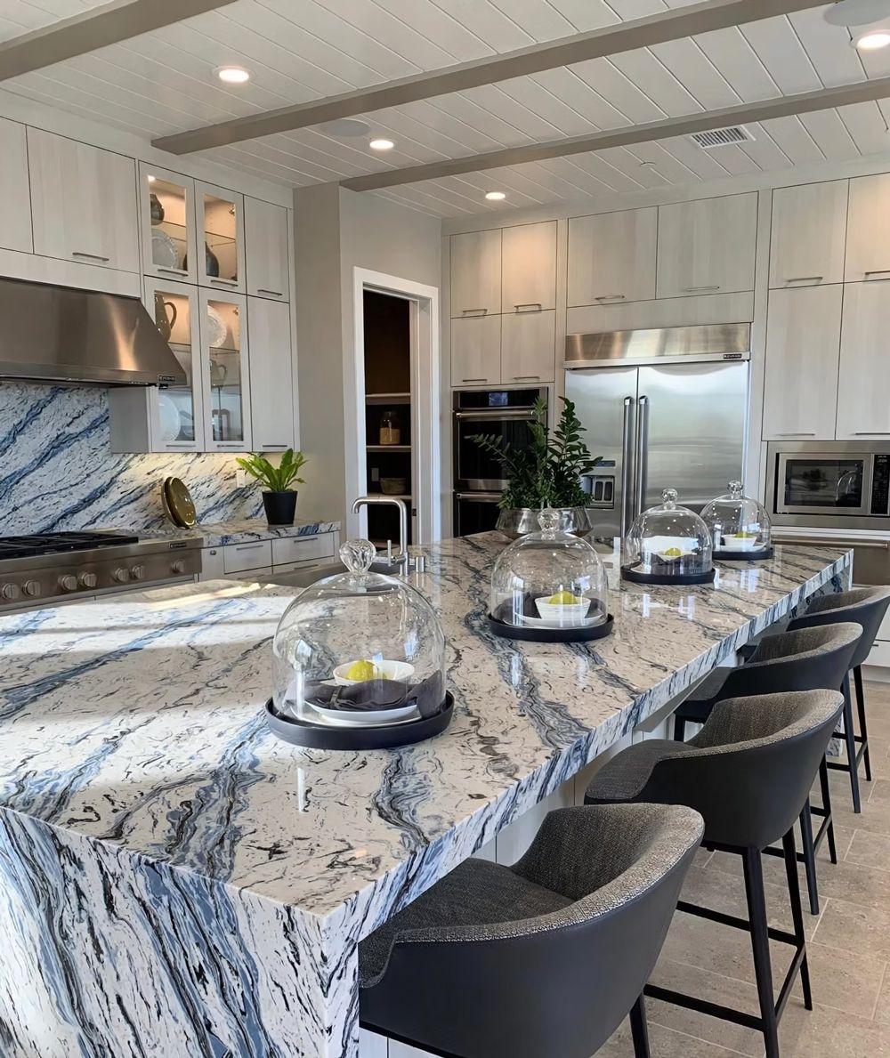 Bright modern kitchen with large marble island, black stools, stainless appliances, and glass display cabinets