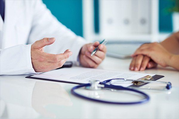 Médico consulta con un paciente en un escritorio; se ven estetoscopio, documentos y manos.