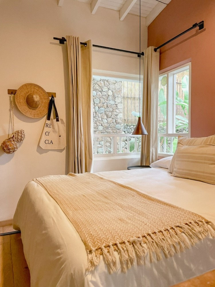 A bedroom with a bed , a window and a straw hat hanging on the wall.