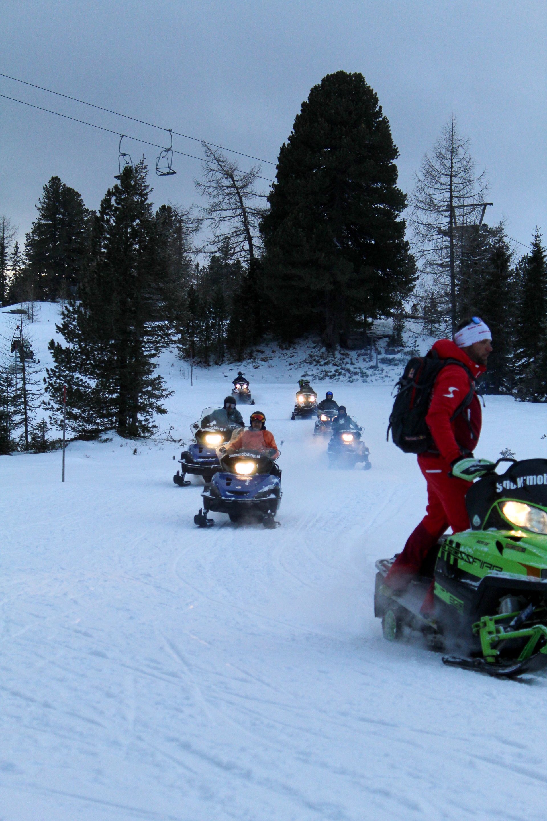 Group of snowmobiles in snowy terrain, led by a person in a red suit. Ski lift visible.