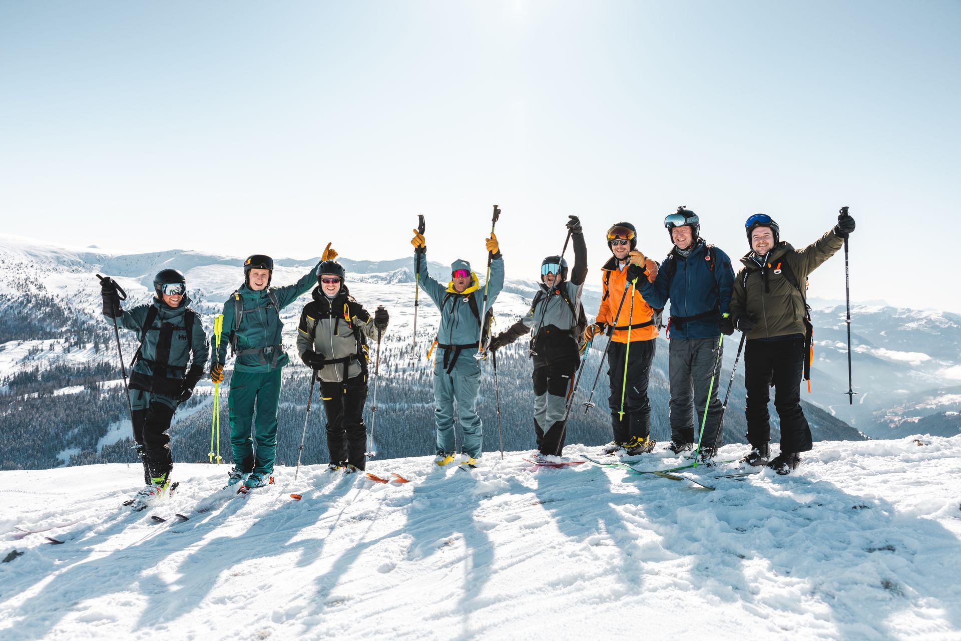 Group of skiers on snowy mountaintop with arms raised in celebration under blue sky.