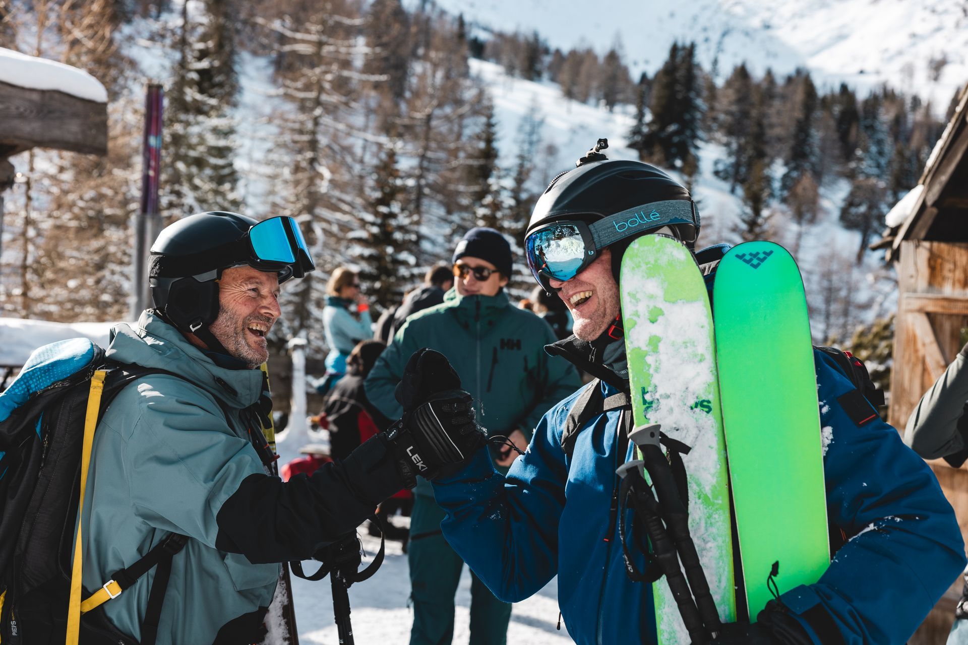 Two skiers, in helmets, laughing outdoors near a snowy mountain. One holds bright green skis.
