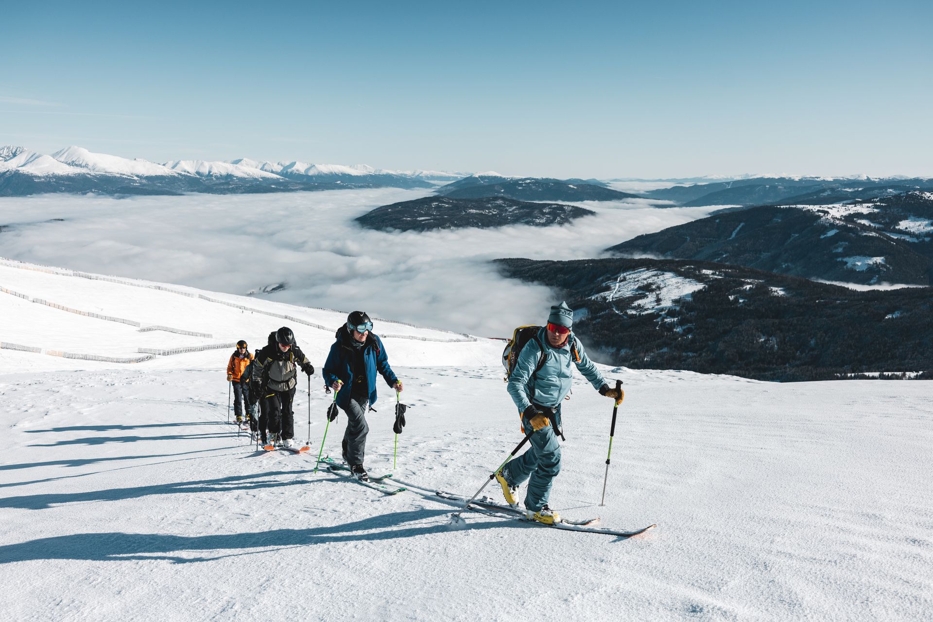People ski touring on a snow-covered slope, above a layer of clouds, under a blue sky.