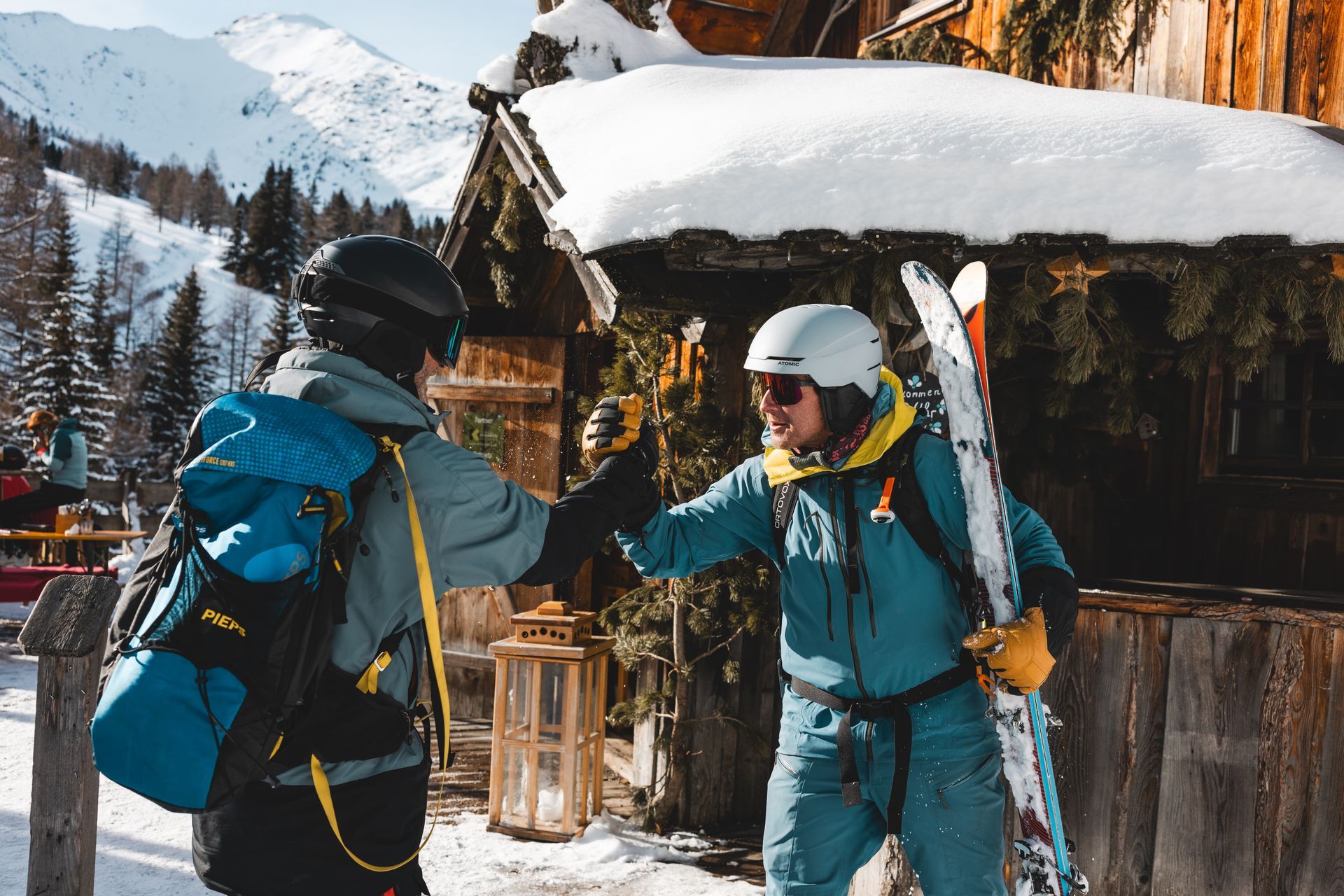 Two skiers bump fists outside a snow-covered cabin in the mountains.