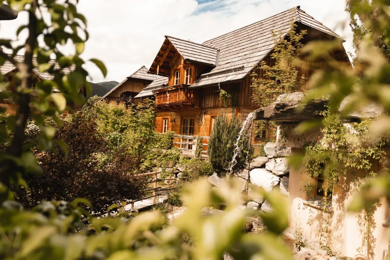 Wooden house nestled in lush greenery, with a balcony and stone wall.
