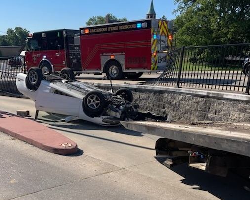 White car overturned near a bridge with a fire truck in the background, likely after a crash.