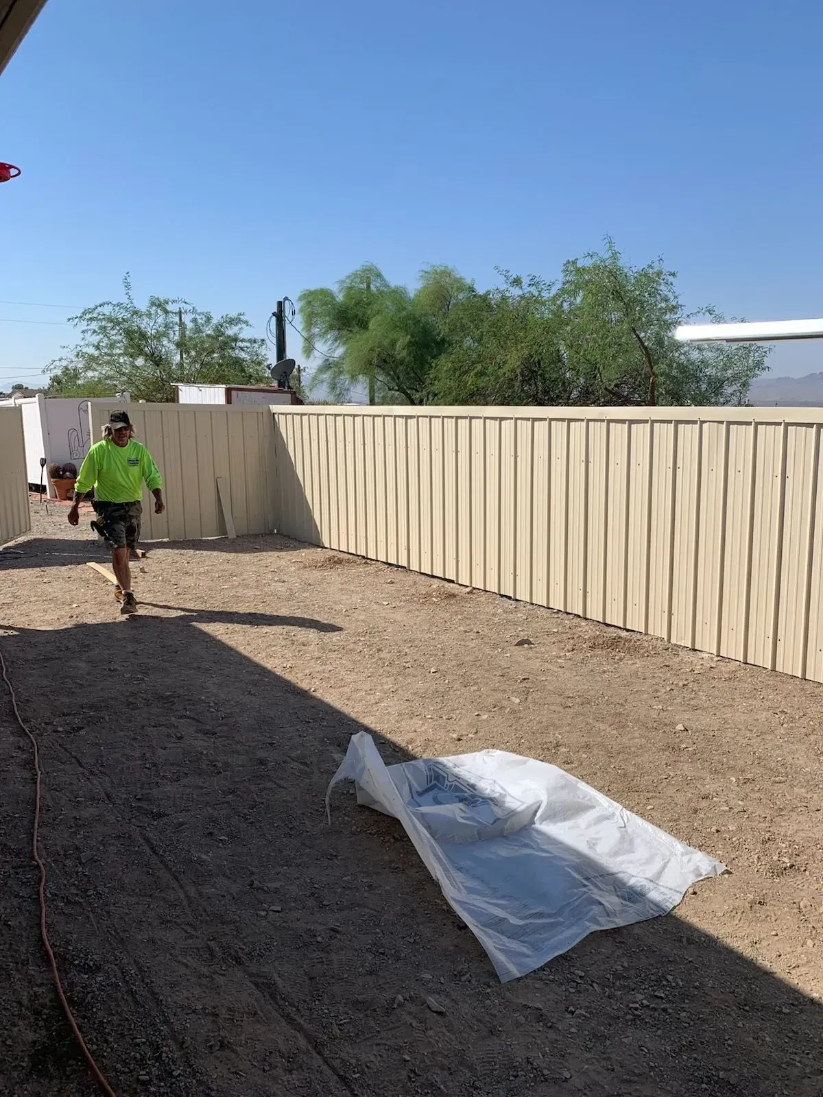 Person walking beside a beige fence in a sunny dirt yard with a white tarp on the ground