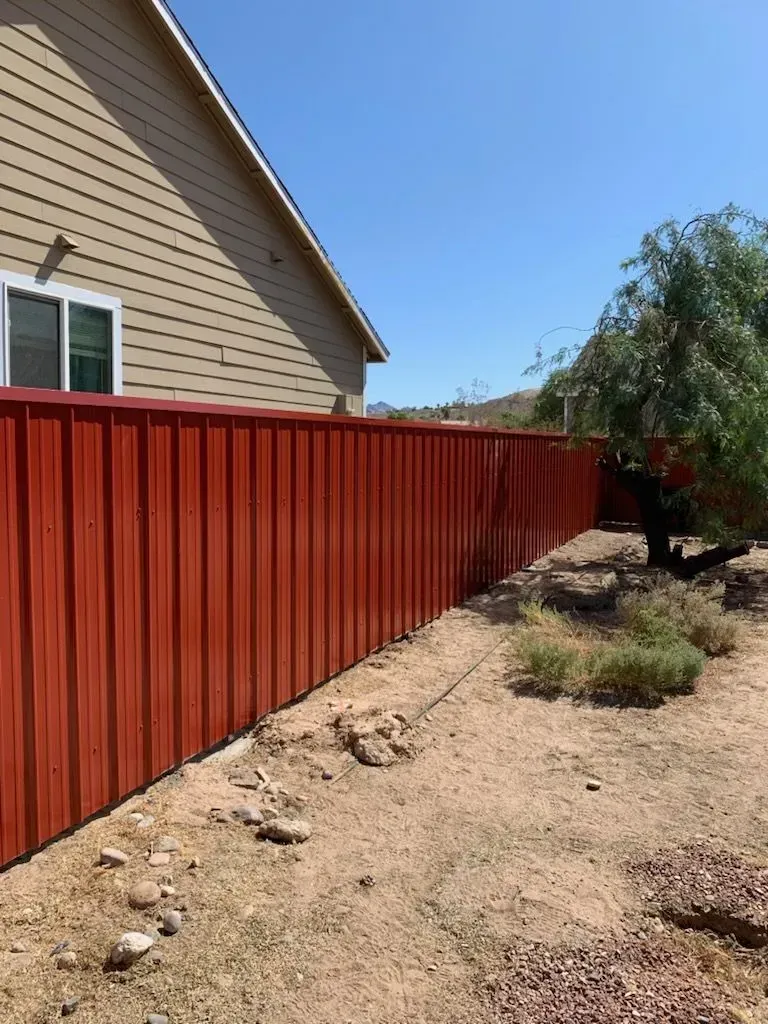 Red wooden fence beside a tan house along a dry gravel yard with a tree.