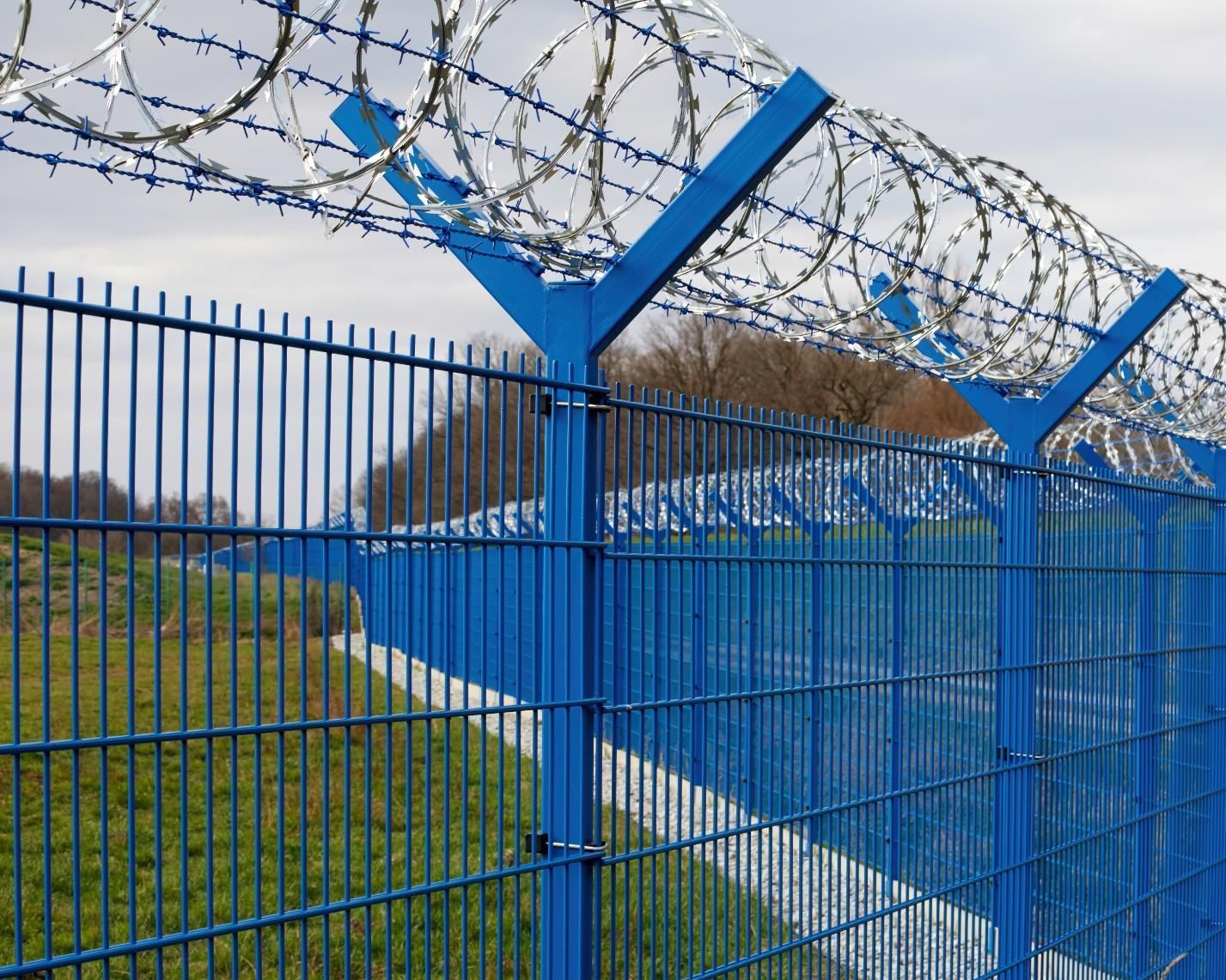 Blue barbed-wire fence along a paved path beside a grassy field under a cloudy sky