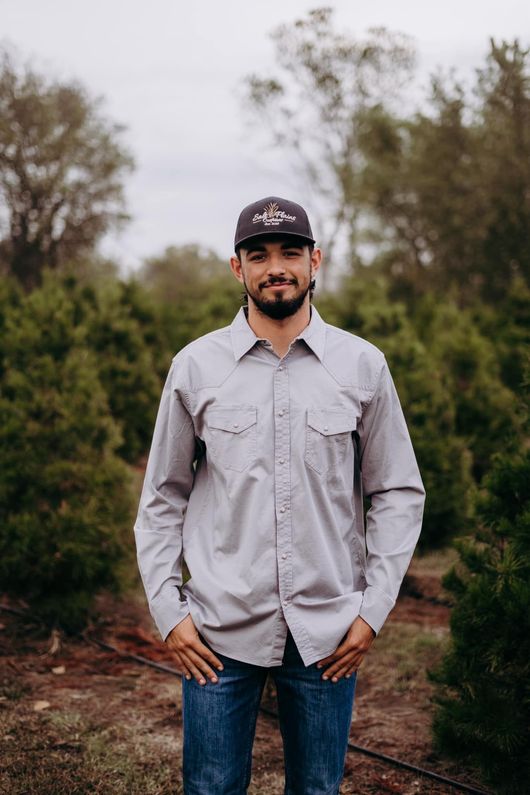 Man standing outdoors on a dirt path, wearing a gray shirt, blue jeans, and a black cap.