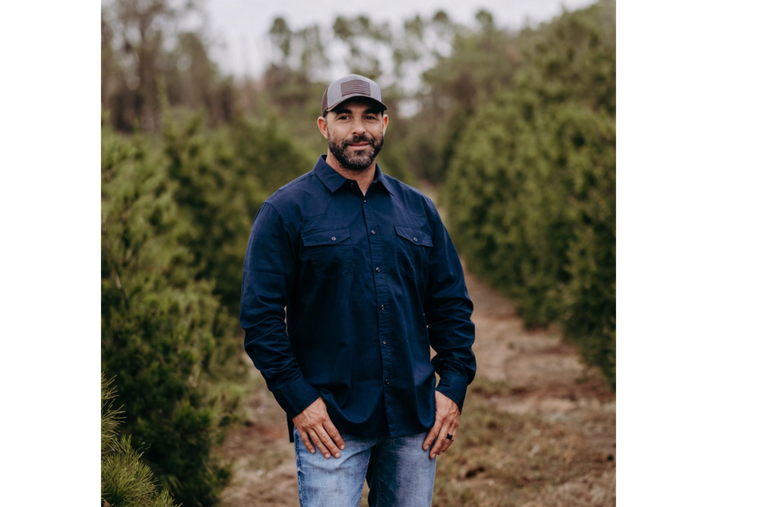 Man in a navy shirt and cap standing on a dirt path between rows of tall green bushes.