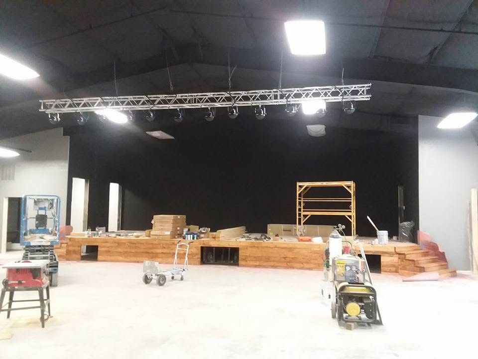 Indoor stage under construction with truss lighting, stacked wood, ladders, and equipment on a black backdrop
