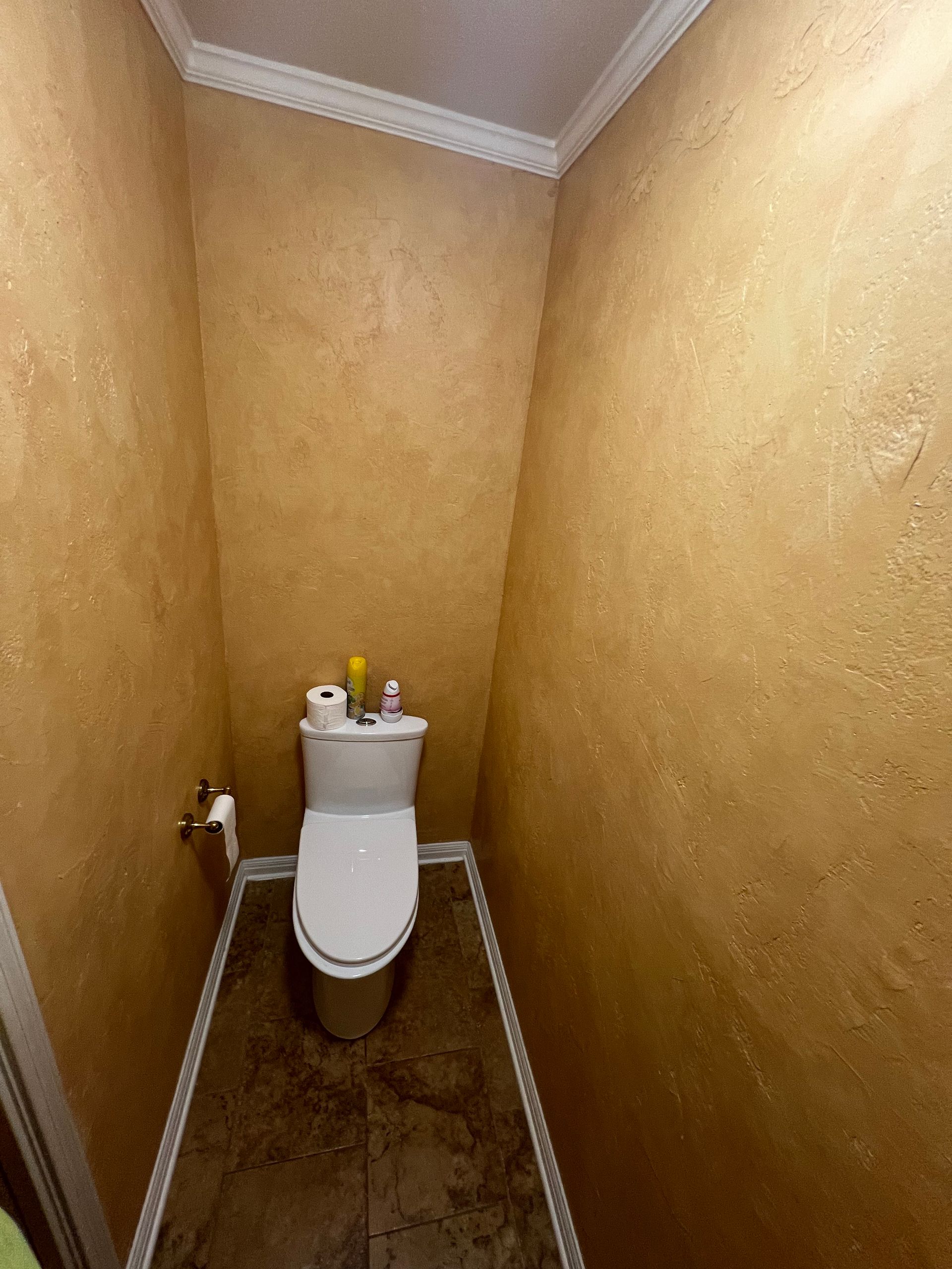 Narrow beige restroom with a toilet centered at the far end and worn walls and floor