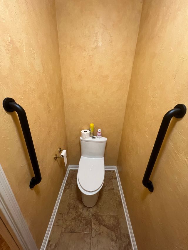 Small restroom with a white toilet, tan walls, and black grab bars on both sides.