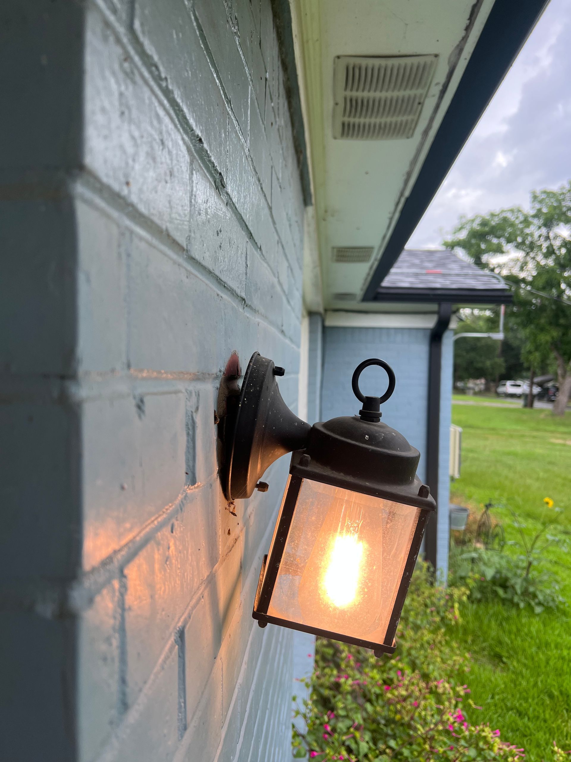 Lit black lantern-style wall sconce mounted on a white house exterior beside a grassy yard.