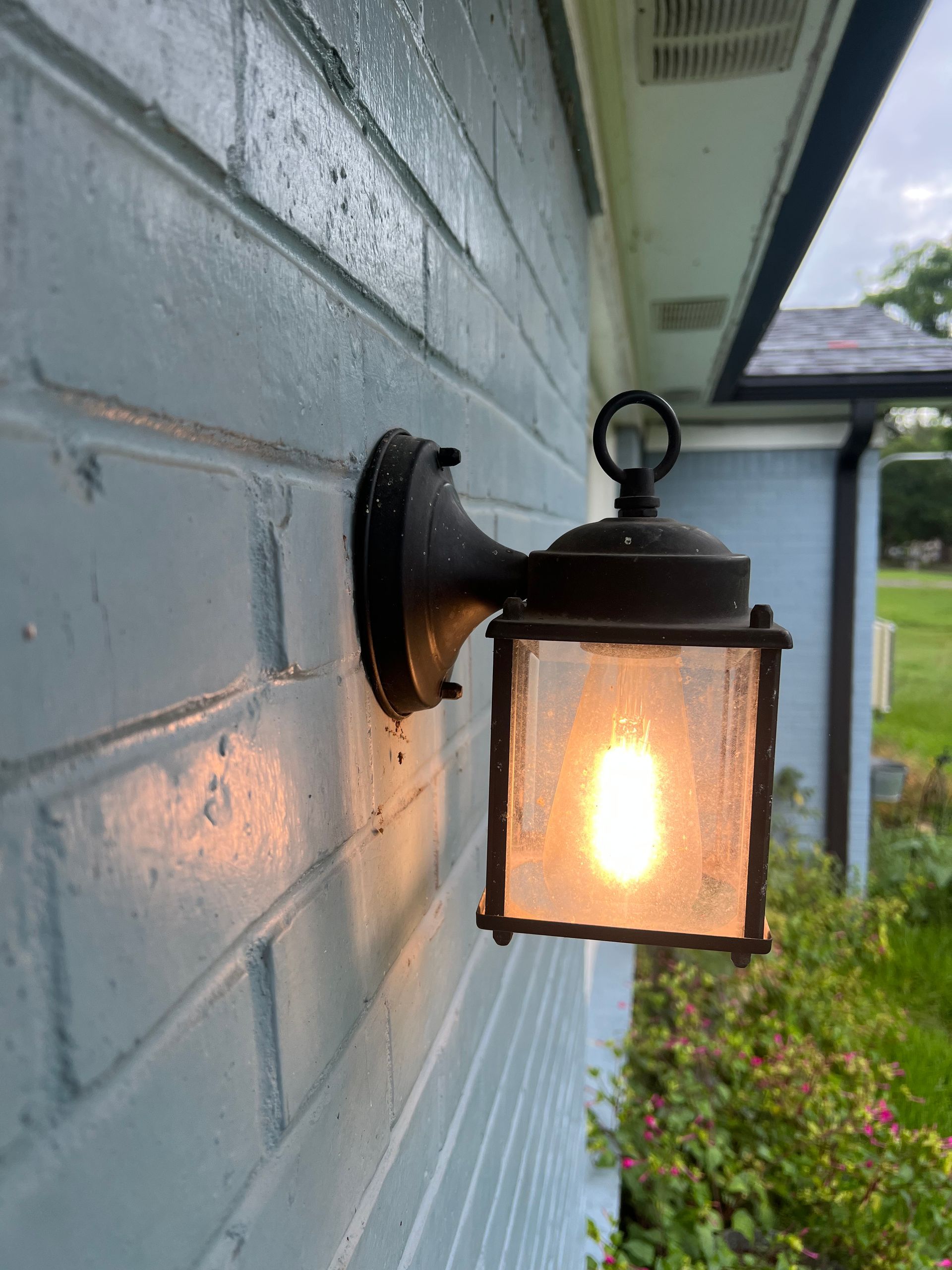 Lit black wall lantern glowing on a blue brick exterior wall beside a walkway.