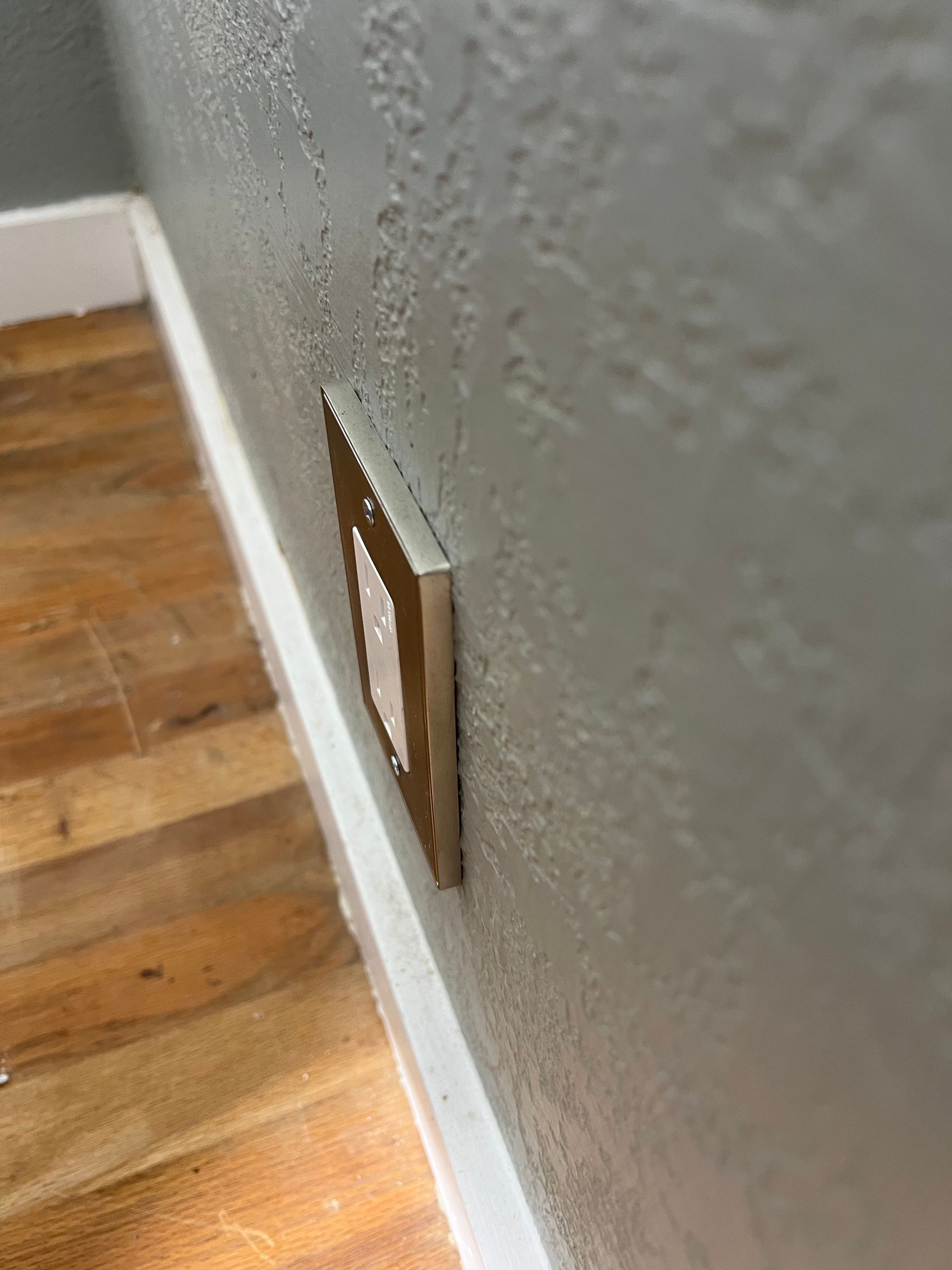 Wall outlet on a textured gray wall beside a hardwood floor and white baseboard.