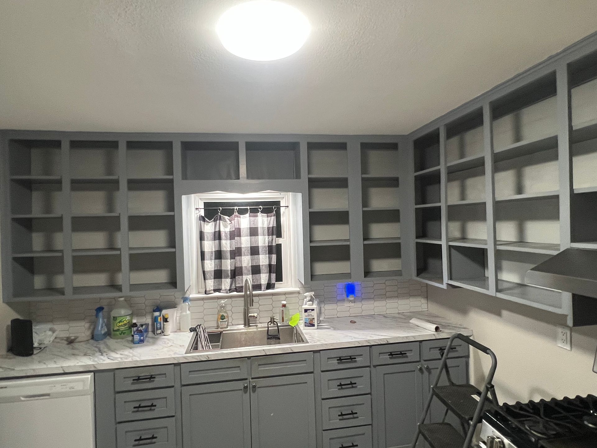 Gray kitchen with unfinished cabinets, countertop clutter, and a window with a checkered curtain