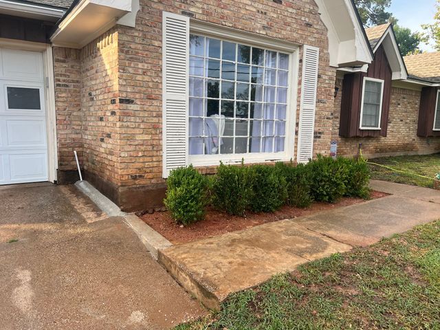 Brick house exterior with large front window, white shutters, and shrubs along the walkway