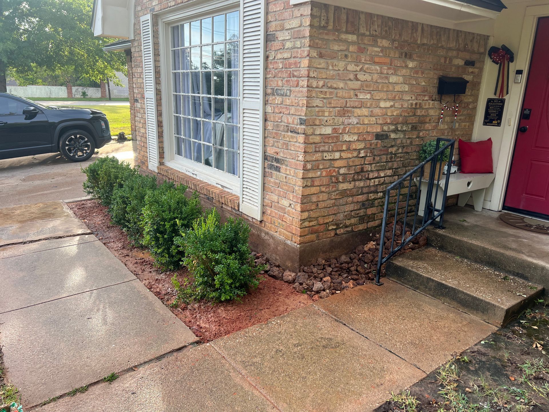 Brick house corner with front steps, red door, window, and shrubs beside a wet walkway