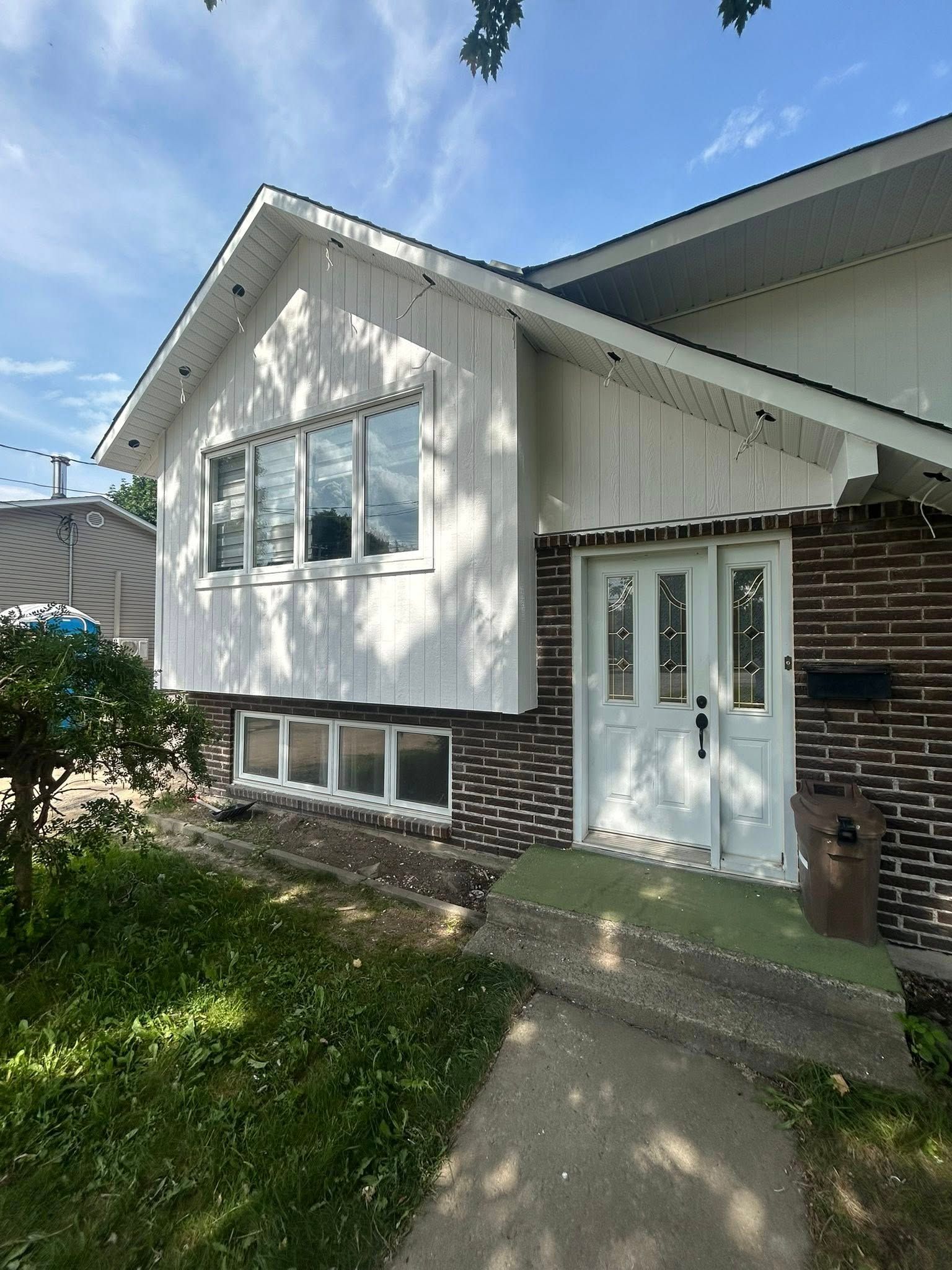 Maison blanche avec façade en briques et porte d'entrée. Pelouse verte au premier plan sous un ciel bleu.