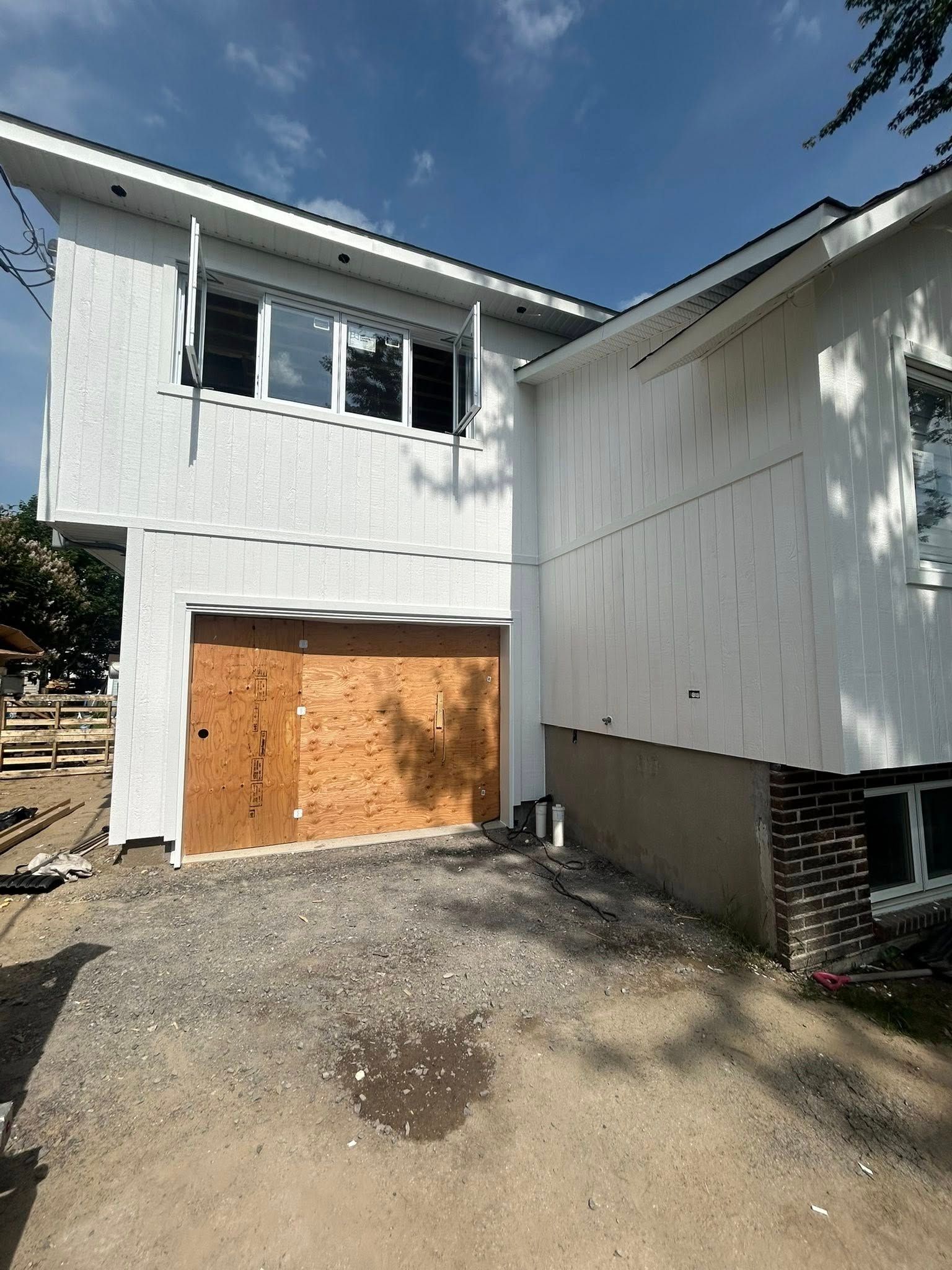 Extérieur d'une maison en construction ; bardage blanc, porte de garage en contreplaqué, allée en gravier.