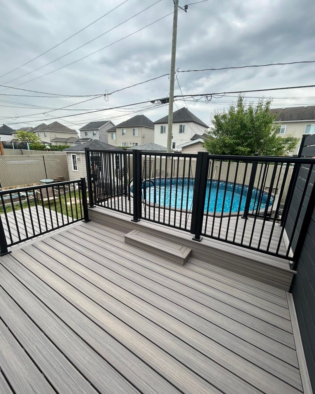 Une terrasse en composite avec une rambarde noire, donnant sur une petite piscine hors sol et des maisons de banlieue par temps nuageux.