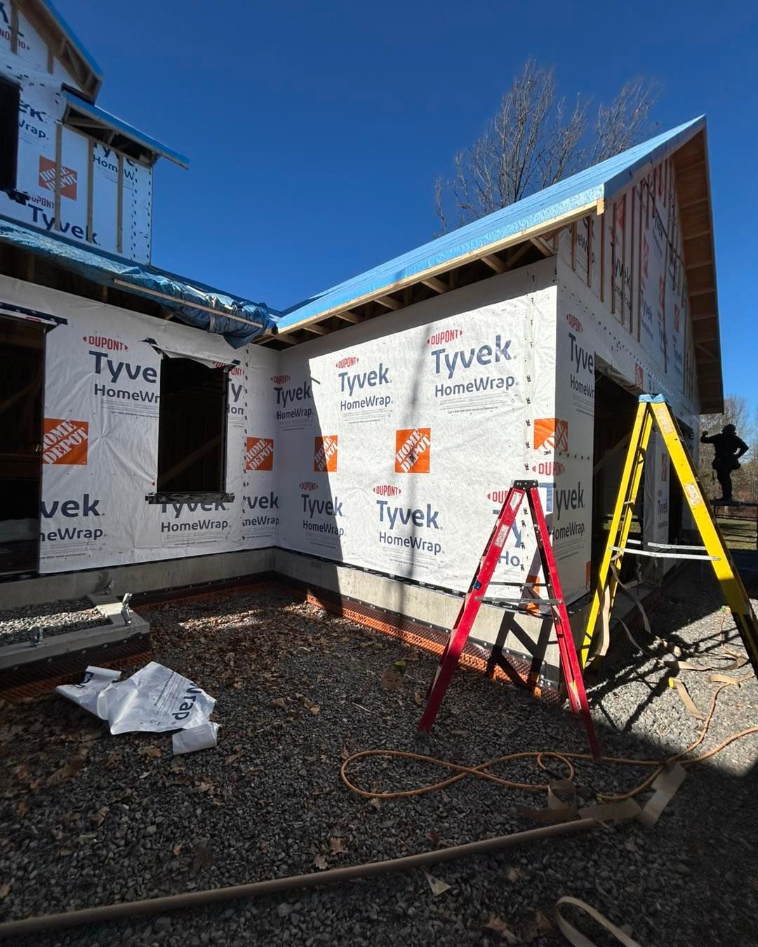 Bâtiment en construction, vue extérieure. La structure est recouverte d'une bâche Tyvek ; ciel bleu au-dessus. Échelle en A et échelle rouge visibles.