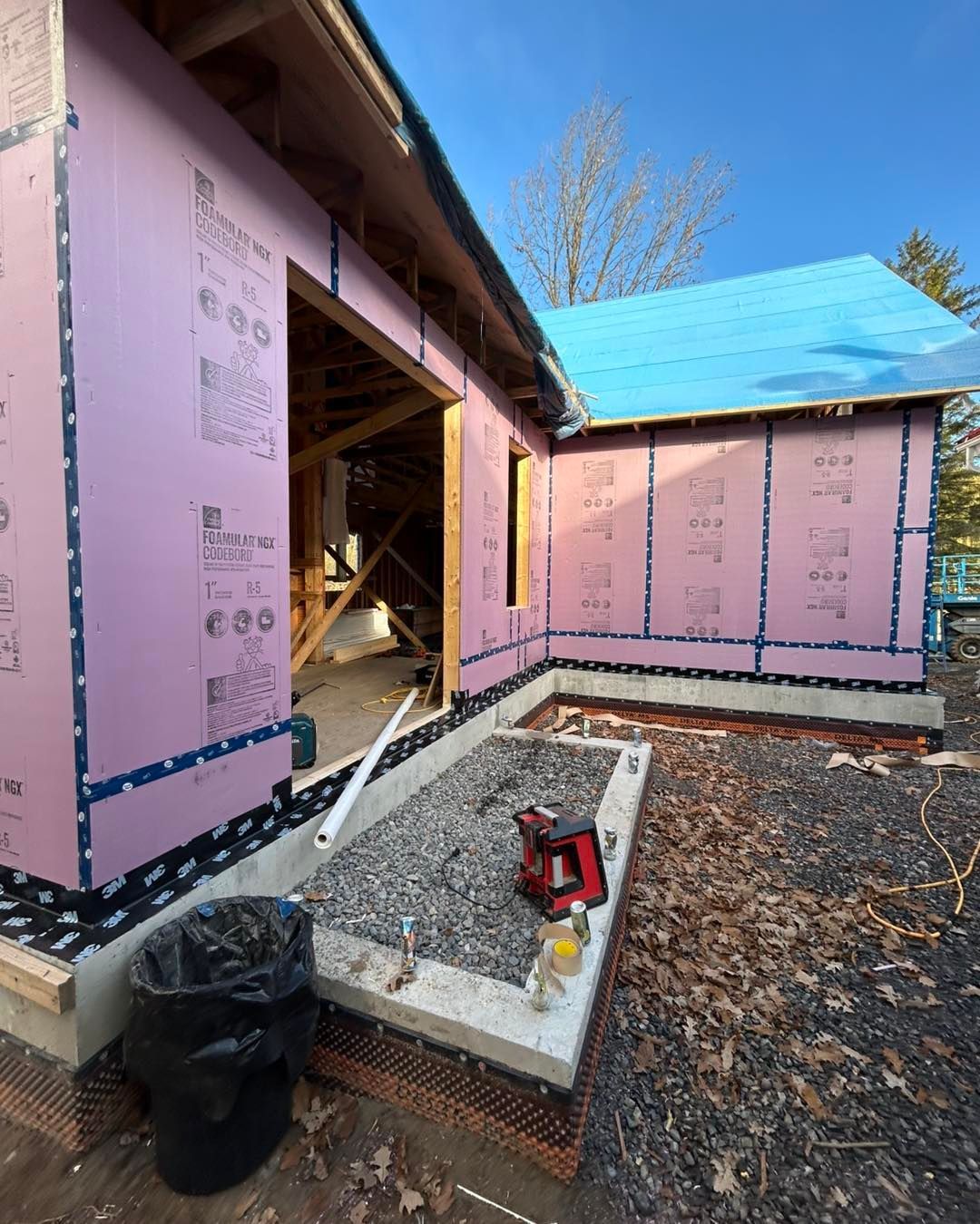 Chantier de construction, extérieur d'un bâtiment avec isolation rose, toit en bâche bleue et base en gravier.