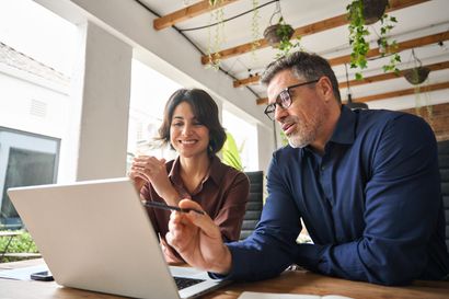 A man and a woman are sitting at a table looking at a laptop computer.