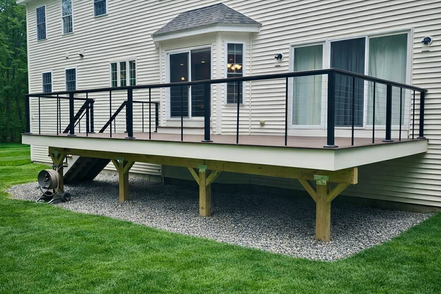 Raised wooden deck with black railing and stairs beside a beige house over gravel and grass