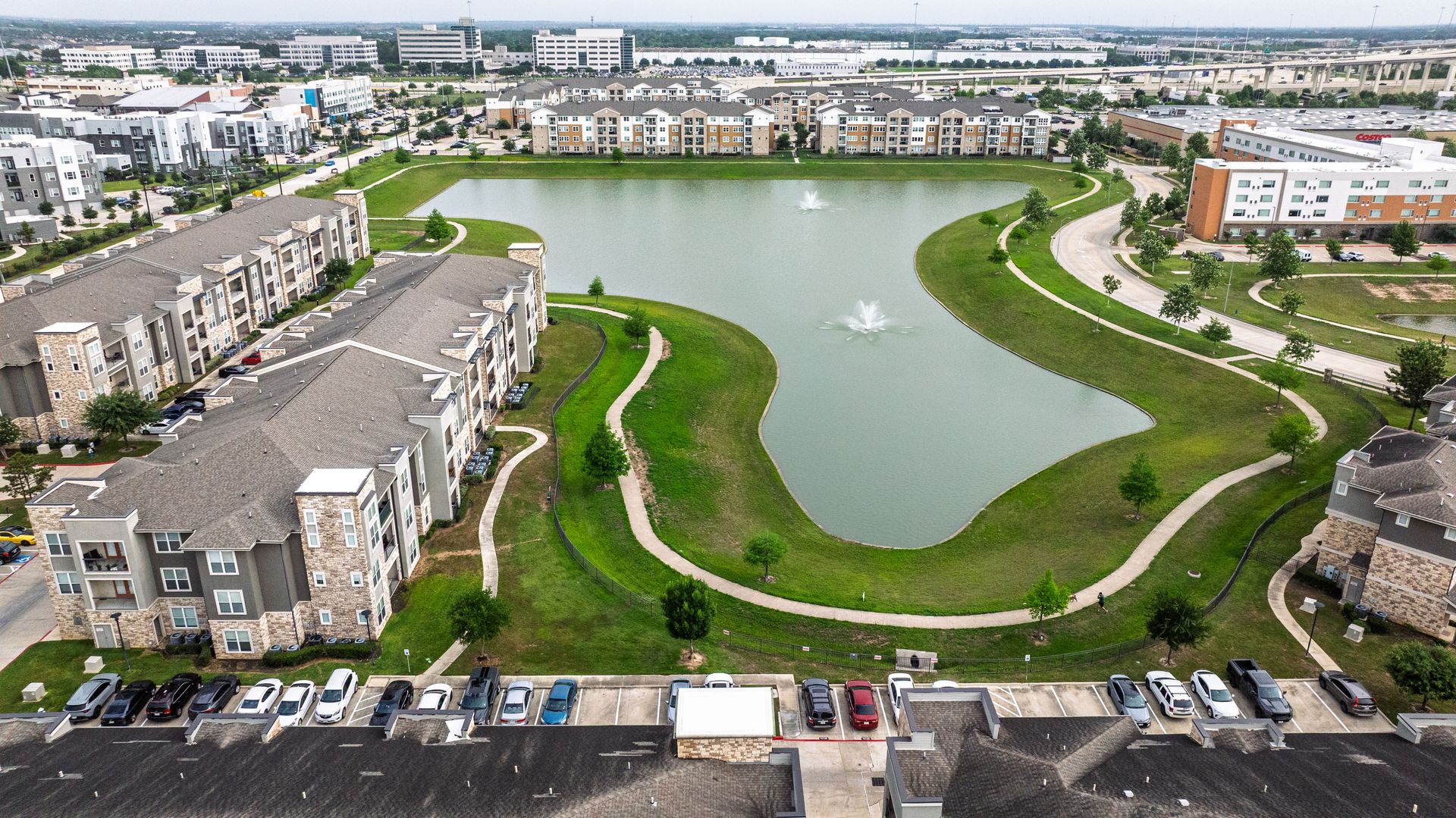 Aerial view of a green lakeside park with apartment buildings and parking lots around it at Olympus Grand Crossing, offering apartments for rent near Houston, TX.