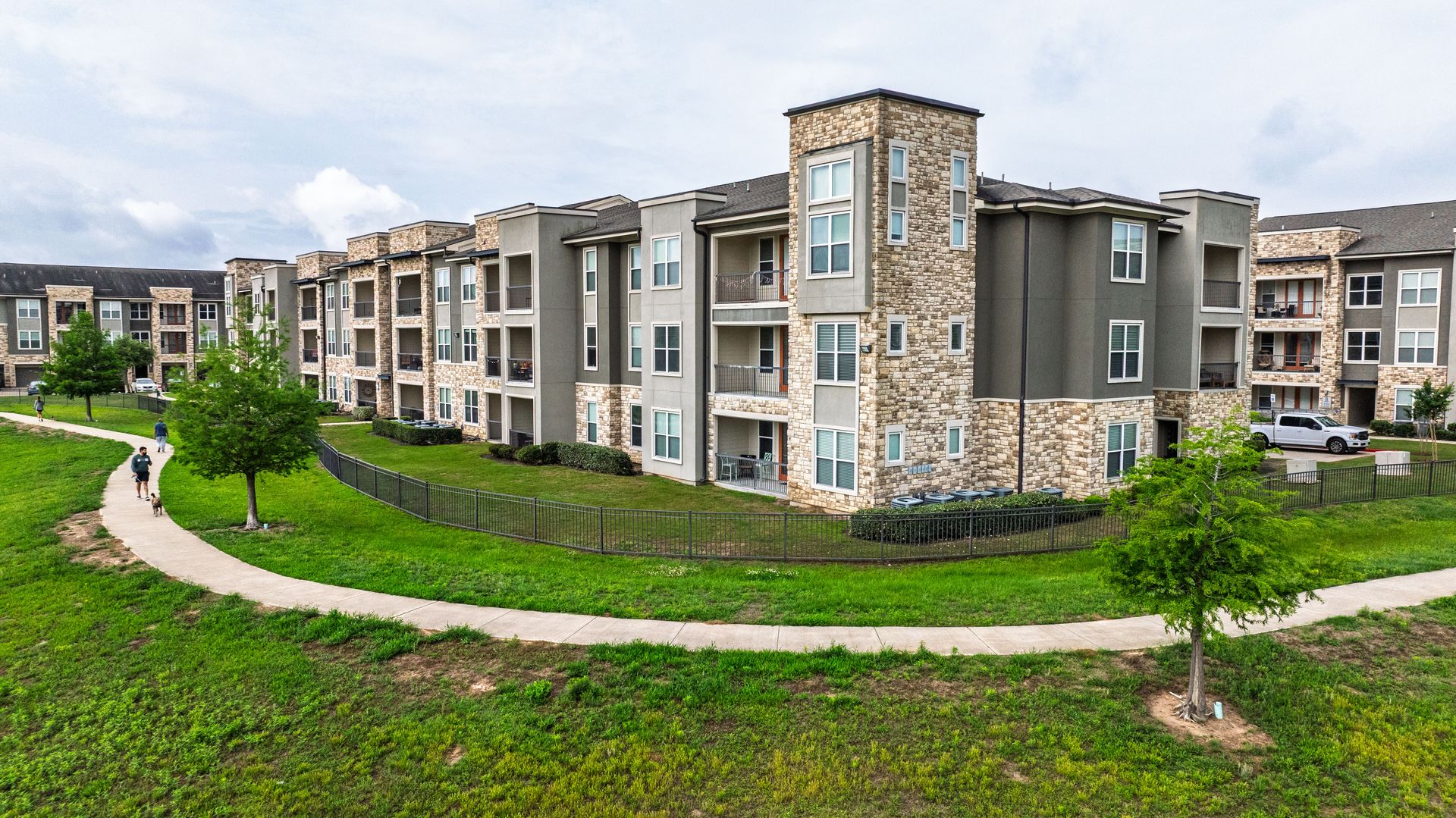 Modern apartment complex with gray stone facades, green lawns, and a winding sidewalk under cloudy skies at Olympus Grand Crossing, offering apartments near Houston, TX.