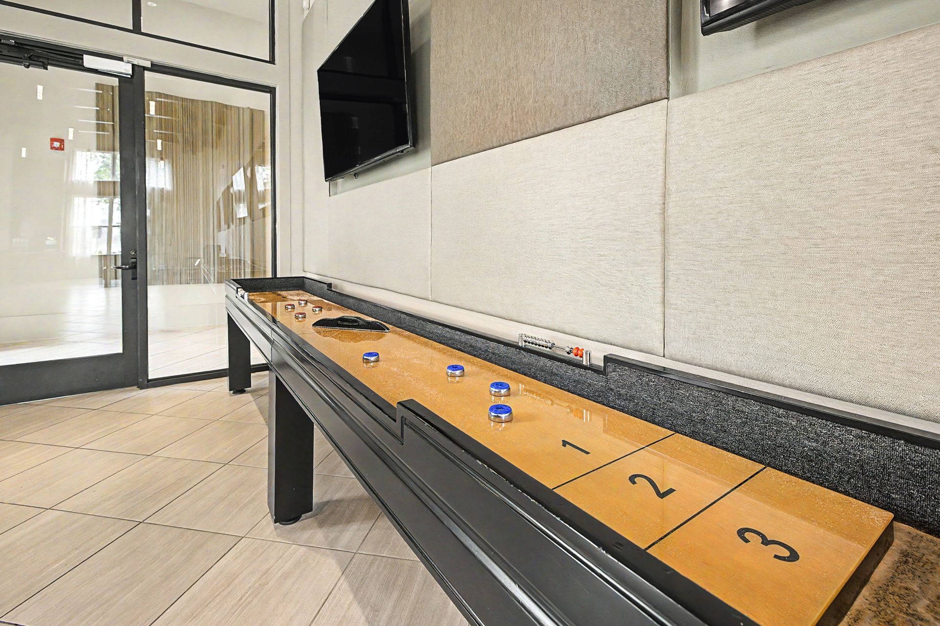 Shuffleboard table in a lobby with numbered lanes and blue pucks near glass doors at Olympus Grand Crossing, offering pet-friendly apartments in Katy, TX.