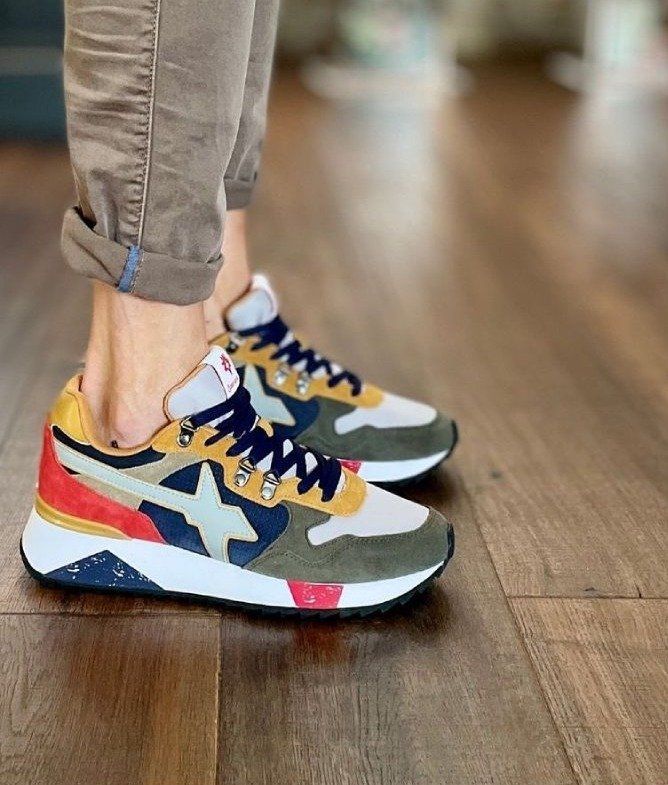 Persona che indossa scarpe da ginnastica colorate, con dettagli verde oliva, blu, rosso e oro. Pantaloni color cuoio arrotolati su un pavimento di legno.