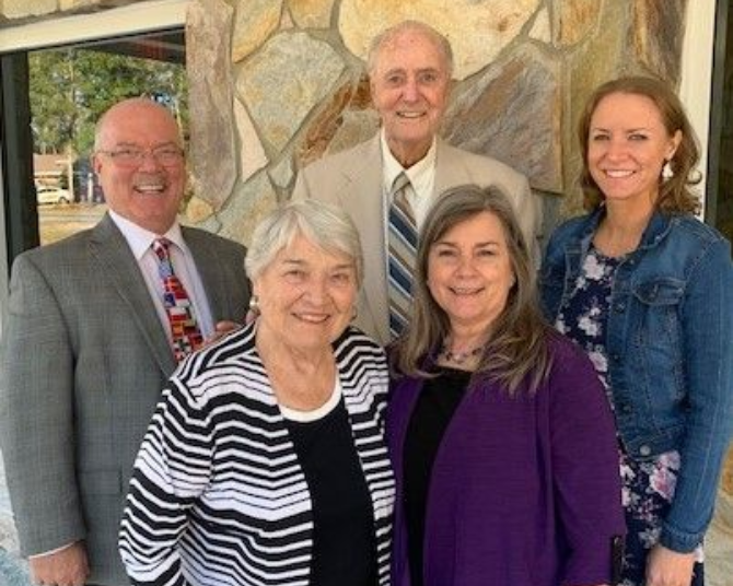Five smiling people standing indoors in front of a stone wall, dressed in business attire.